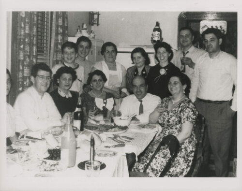 A large group of people posing for a photo around a dinner table. Several are holding bottles.