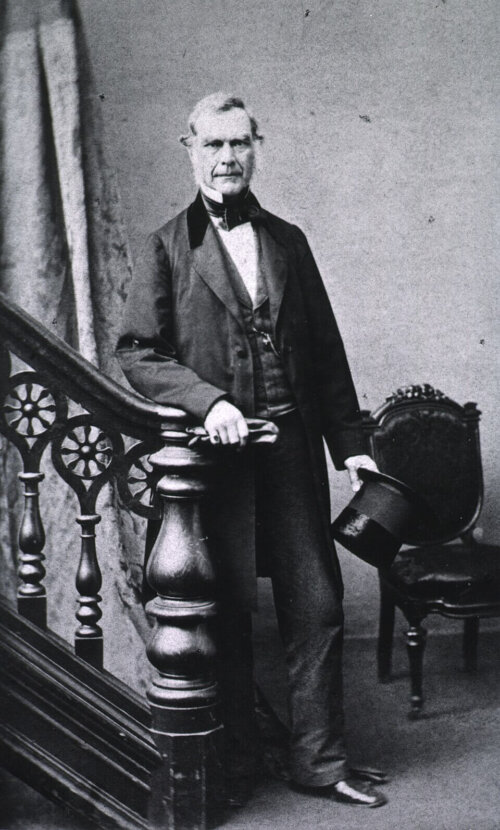 A man with mutton chops standing at the bottom of a staircase and holding a top hat.