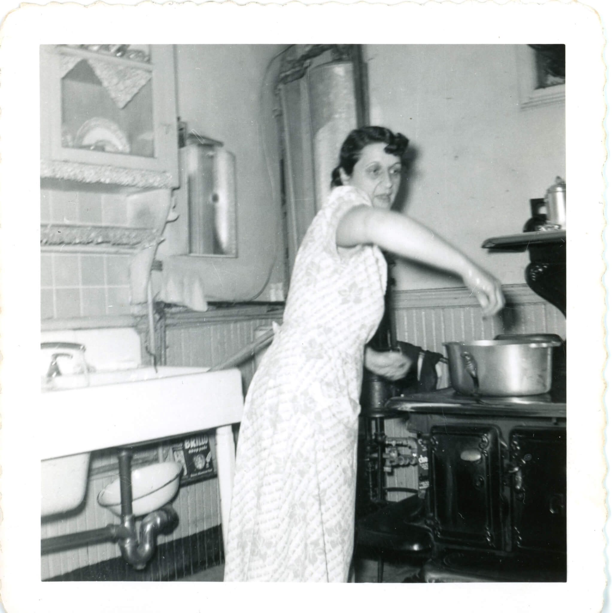 Black and white image of a woman standing between a porcelain sink and a metal stove holding her hand over a pot.
