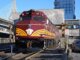 A red and gold train engine pulling out of a station. There is a car parked to the right and a bridge in the background on the left.