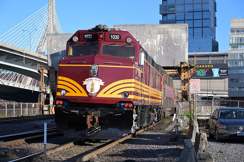 A red and gold train engine pulling out of a station. There is a car parked to the right and a bridge in the background on the left.