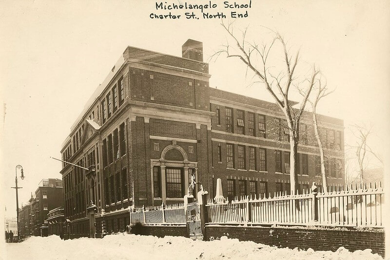 A school building next to a cemetery. The ground and sidewalk is covered in snow.