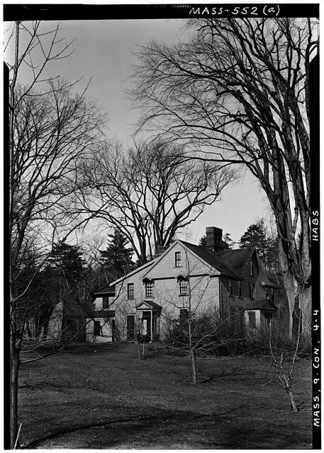 A black and white photo of a three story farm house in an area of trees.