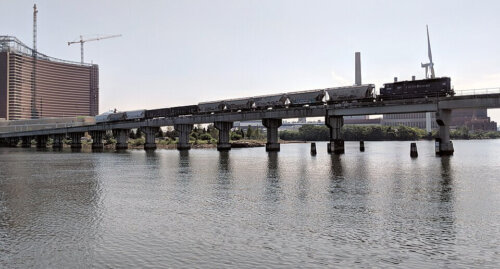 A train with several freight cars crossing a train bridge over the river with a large building under construction in the background.