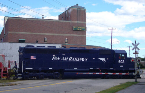 A blue train engine crossing a road with a brick building in the background.