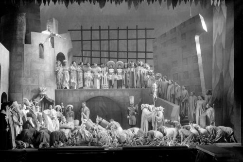 A large group of actors on a stage bowing down to a figure on the left side of the stage while a man at the top of a castle set points a sword down at a severed head on a stick.