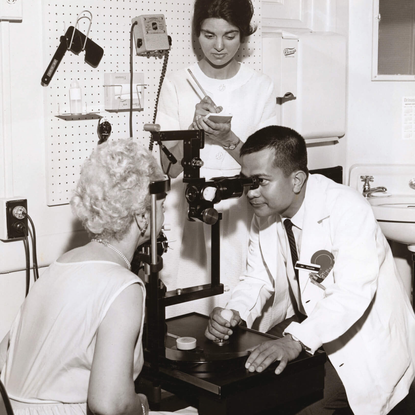 A man in a white coat looks into a device to examine and eye while a woman in a nurses uniform takes notes.