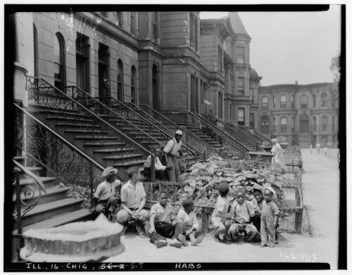 A group of Black children sitting in front of a front garden at the base of a set of brownstones set back from the street.