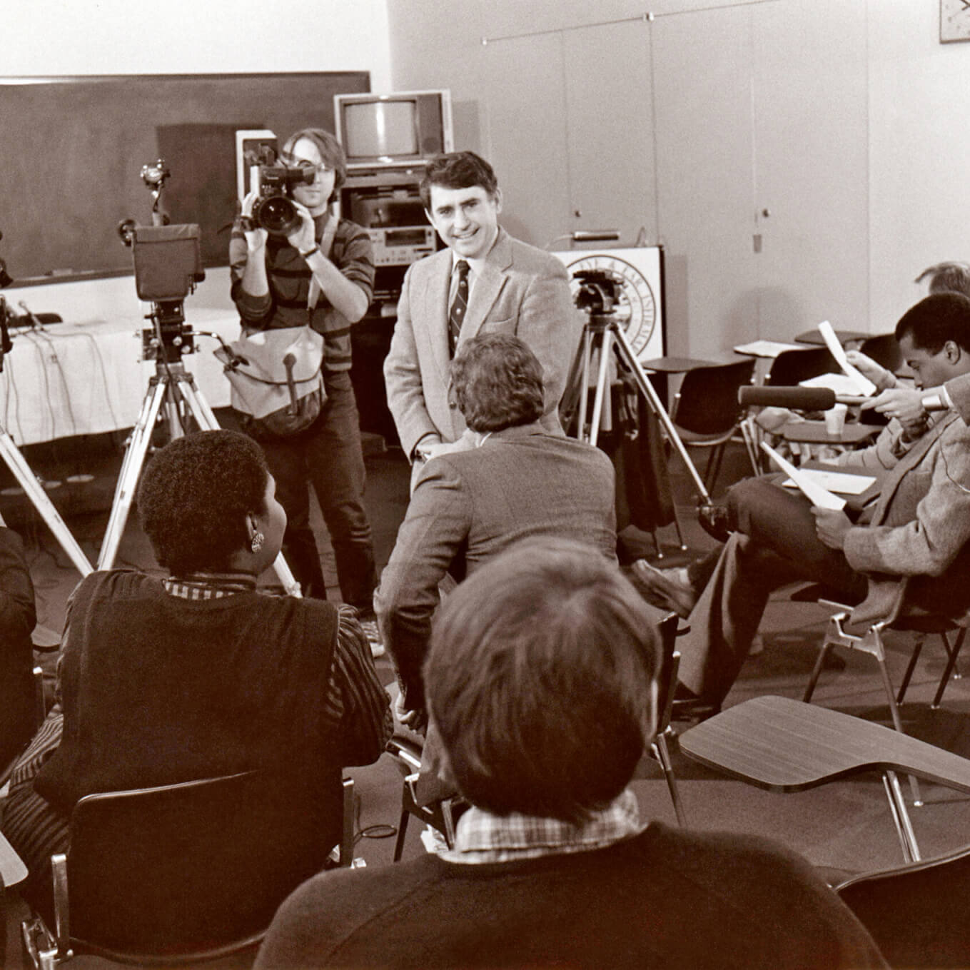 A man surrounded by journalists sitting in a classroom with several cameras pointed at him.