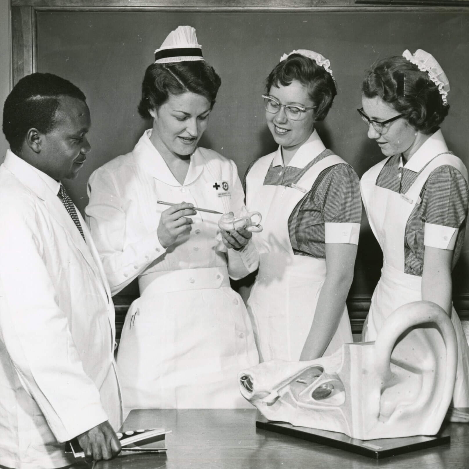 Three women in nurses uniforms and a doctor look at a model of the human ear.
