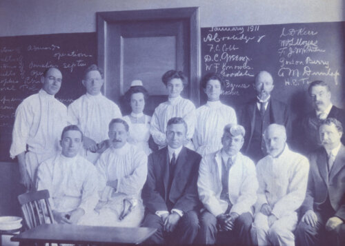 Two rows of doctors and nurses pose for a portrait. There are three women in the center and several of the men wear surgical gowns or suits.