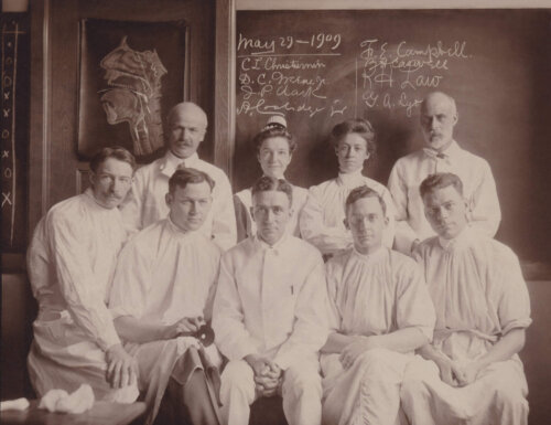 A group of doctors pose for a portrait in front of a blackboard. There are two rows of men in surgical gowns and two women.