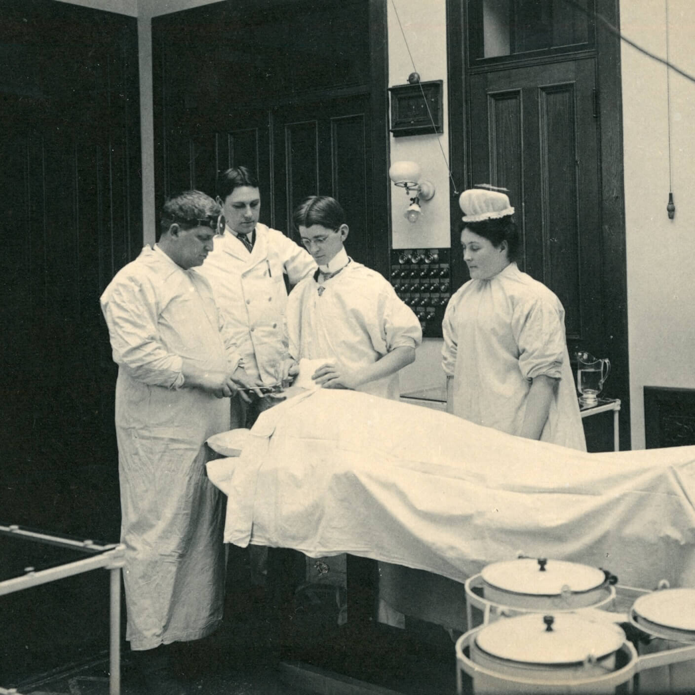Three men and a woman standing around a body covered with a sheet doing medical training.